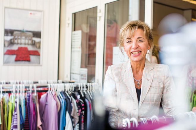 Foto: Eine Mitarbeiterin des Rotkreuz-Shops steht zwischen mehreren Ständern mit farbenfroher Kleidung. Sie blickt lachend in die Kamera.
