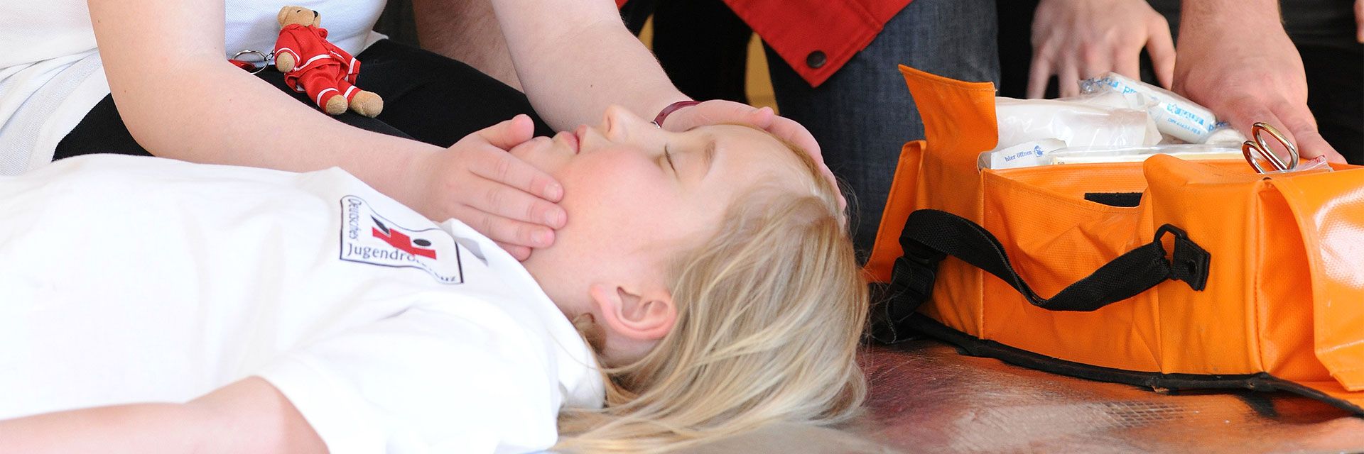 Foto: Bei einer Erste-Hilfe-Übung liegt ein Kind bewusstlos am Boden. Ein anderes Kind überprüft gerade die Atmung. Am Rande des Fotos ist eine Tasche mit Verbandsmaterial zu sehen.