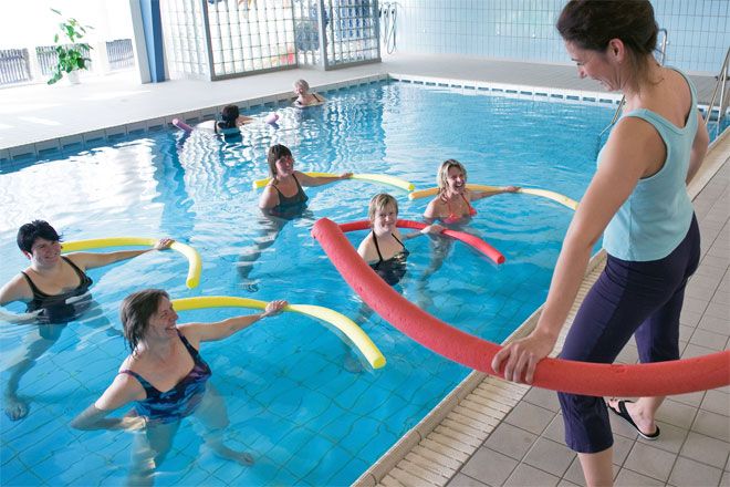 Foto: Mehrere Frauen befinden sich mit Schwimmnudeln in einem Schwimmbecken und blicken die Kursleiterin an. Sie haben sichtbar Spaß zusammen.